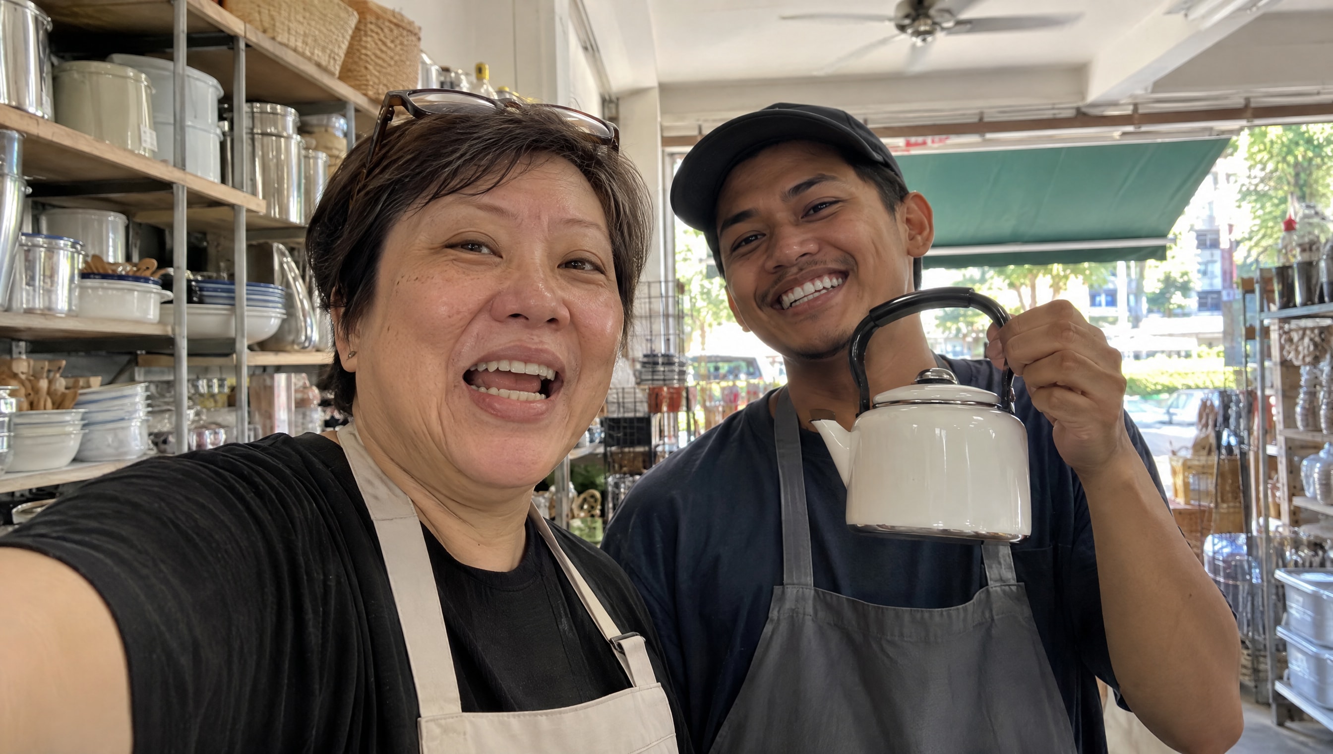 A shop owner films herself on her phone at her shop counter answering a customer's question, while her assistant holds a small product up beside her, both laughing between takes